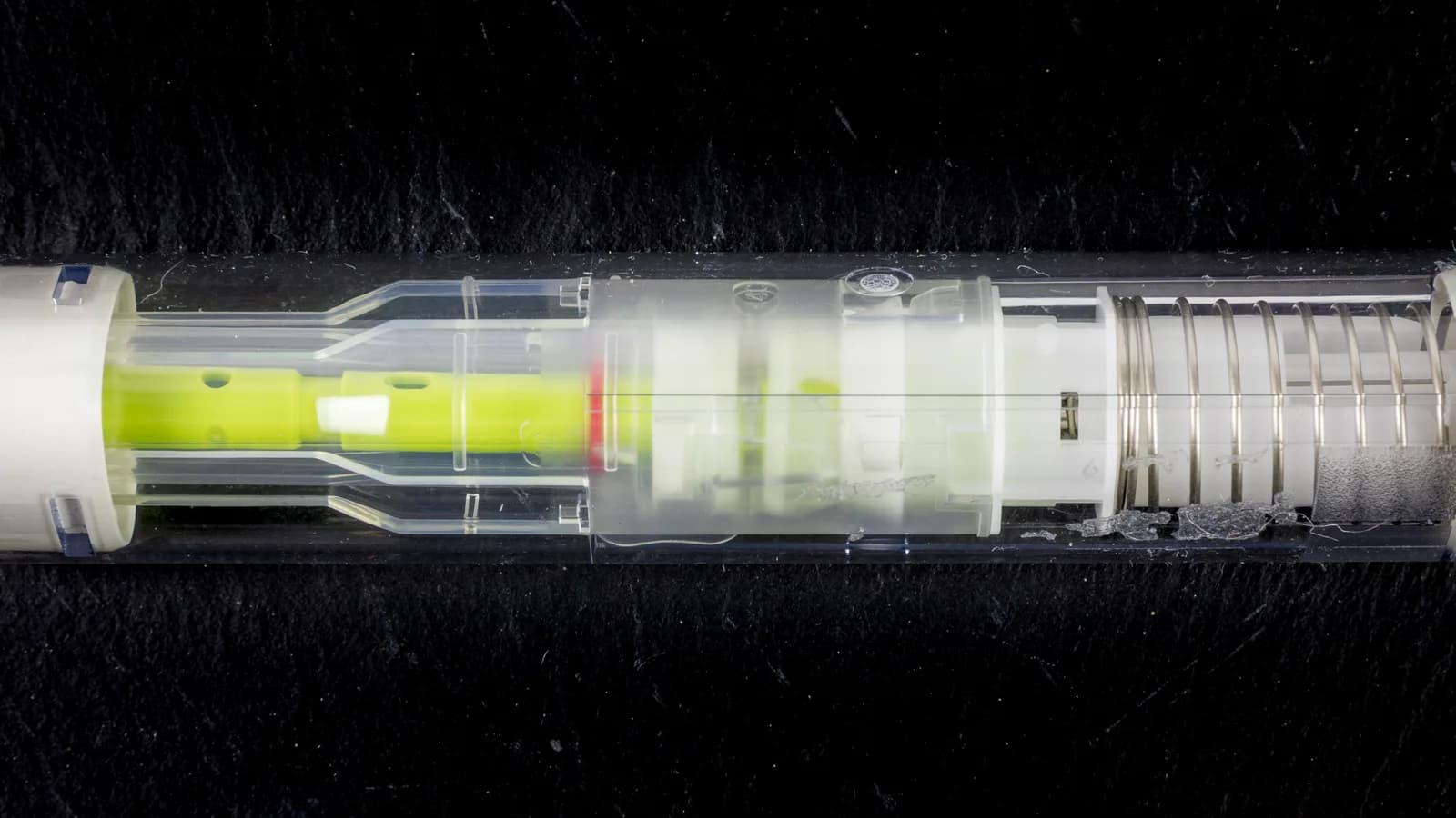 A transparent, unbranded medication autoinjector pen shown in cutaway against a dark background, revealing its spring and plunger mechanism.