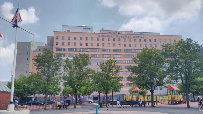The Elmhurst Hospital Center building in Queens seen across a plaza with trees and a flagpole.