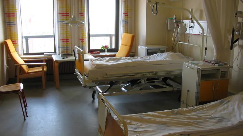 A hospital room with two empty beds, bedside equipment and armchairs by a sunny window.