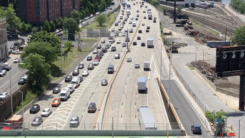 Traffic, including box trucks, on the Major Deegan Expressway seen from the High Bridge, with rail yards beside it and Manhattan in the distance.