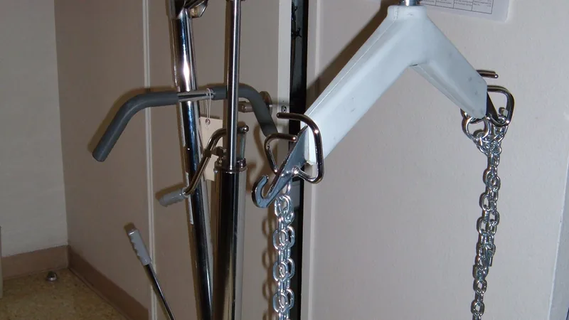 The mast, spreader bar and chains of a mechanical patient lift standing against a wall.