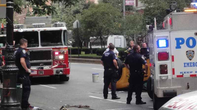 NYPD officers and an FDNY fire truck at a crash scene on a New York City street, with a yellow car tipped on its side between them.