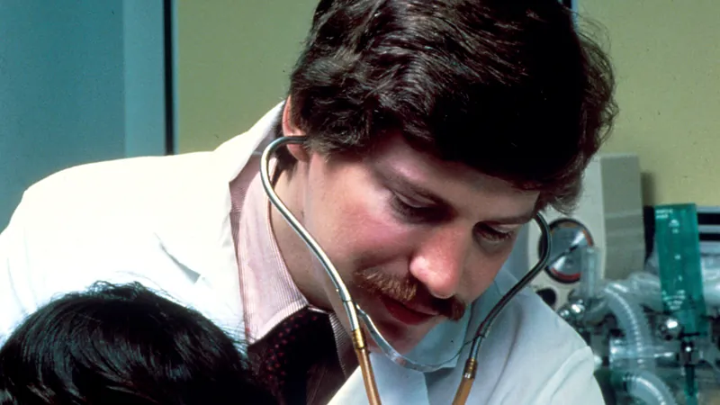 A physician in a white coat listening to a patient’s back with a stethoscope during an examination.