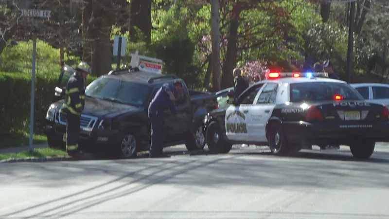 A police officer leaning into a damaged dark SUV while a firefighter stands nearby and a police car with flashing lights blocks the street.