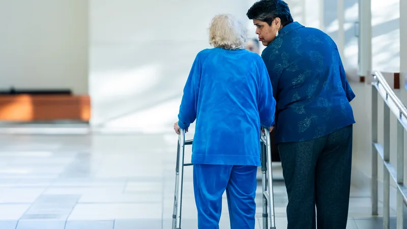 A staff member walks beside an older woman using a walker in a bright corridor, both seen from behind.
