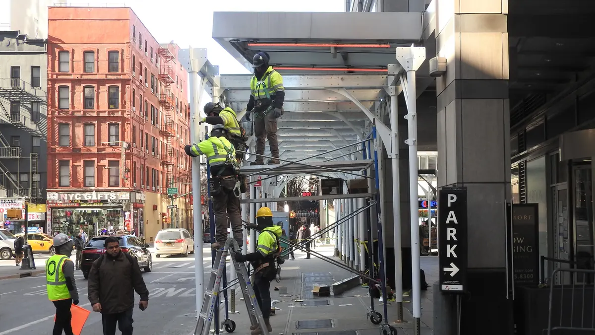 Construction workers in safety vests and hard hats working at height while assembling a sidewalk shed on West 47th Street in Manhattan.