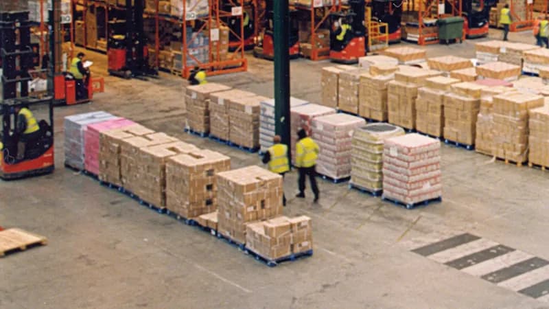 Warehouse workers on foot walking between pallets while reach trucks operate along the racking.
