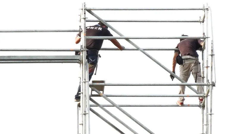 Two workers standing on the upper levels of an open metal frame scaffold without guardrails while assembling it.