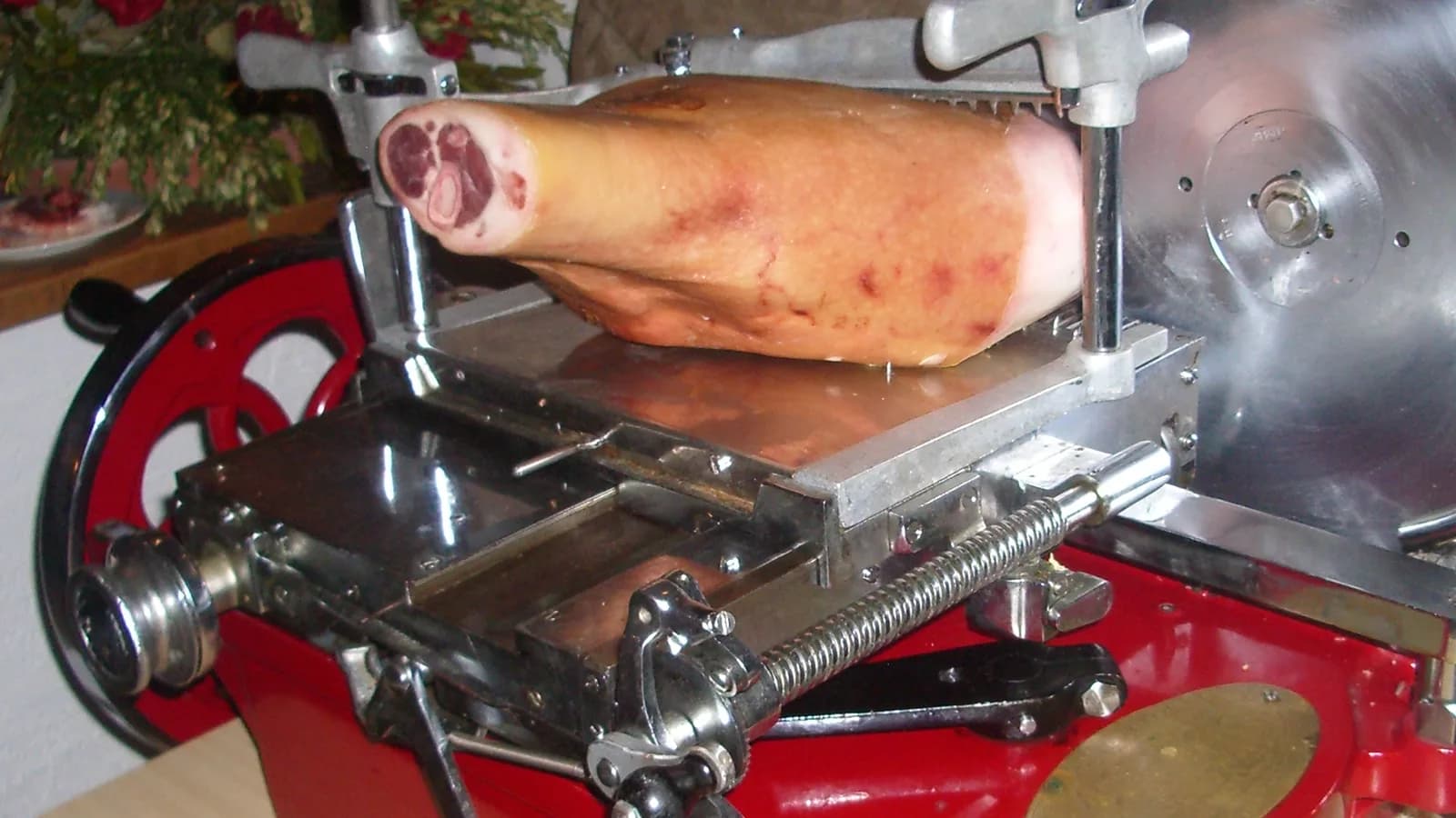 An antique red hand-crank meat slicer holding a ham, with its circular steel blade and slicing carriage in view.