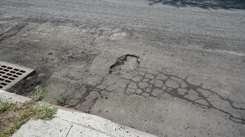 A pothole and cracked asphalt beside a curb and storm drain on a residential street in Queens.