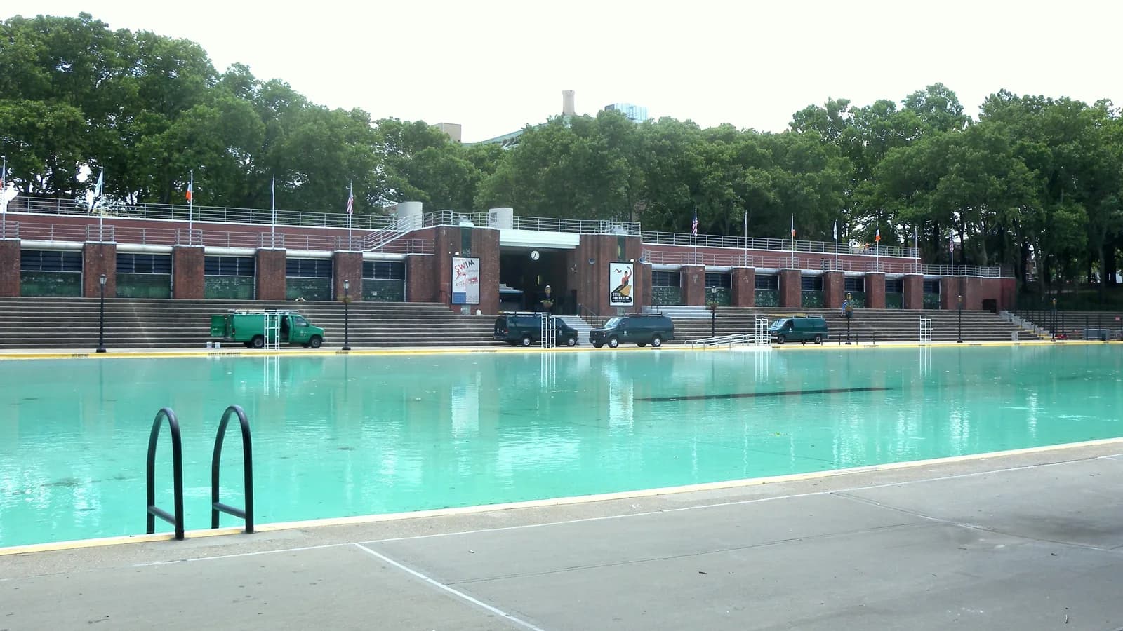 The empty Astoria Pool in Queens before opening day, with an entry ladder in the foreground, a wide concrete pool deck and the bathhouse and bleachers behind it.