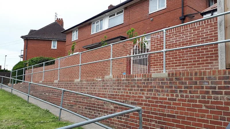 A long access ramp with metal handrails running beside a brick wall and houses.