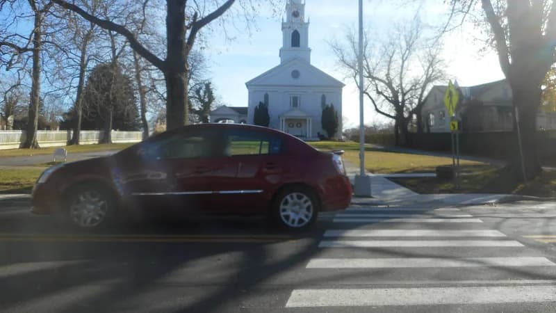 A car passing a marked crosswalk with a pedestrian crossing sign on Main Street in Bridgehampton, with a white church behind.