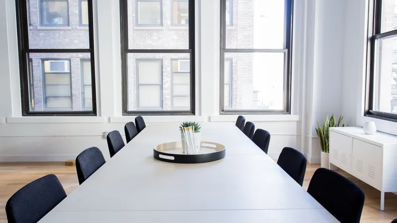 An empty meeting room with a long white table, dark chairs and tall windows in a New York office building.