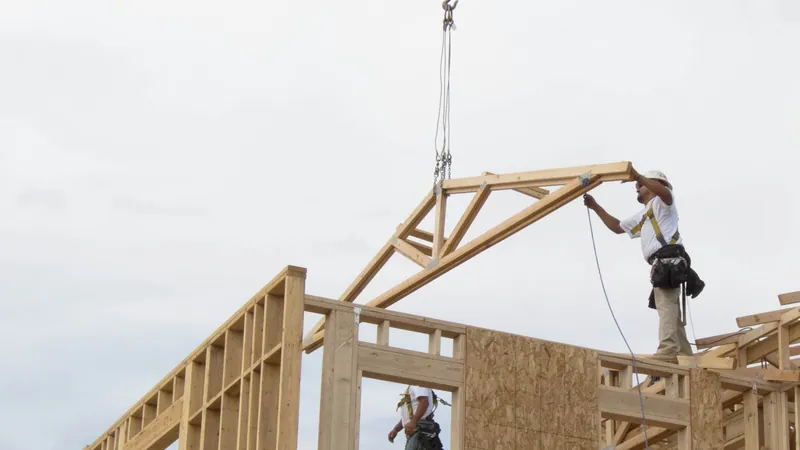 A framing crew member wearing a fall-arrest harness guides a roof truss being lifted onto a house frame.