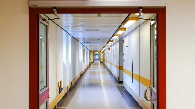 A long, brightly lit hospital corridor seen through an open set of red-framed doors.