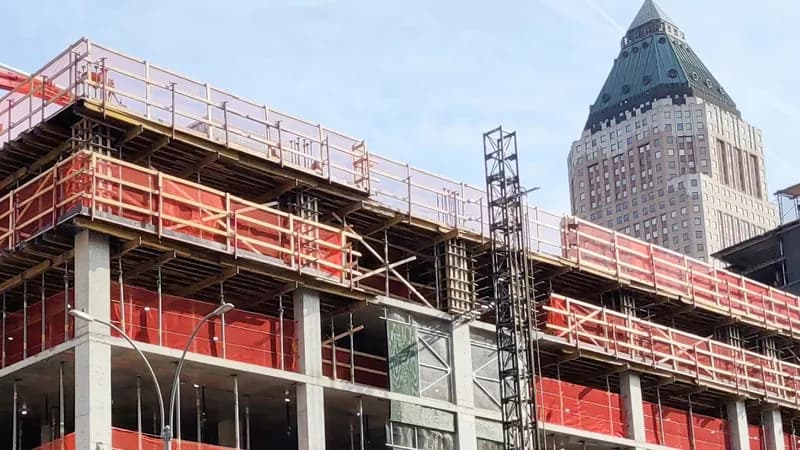 A concrete-frame apartment building under construction in Manhattan, with orange safety netting and a hoist tower.
