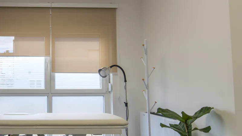 An empty medical examination room with a padded treatment table, an exam lamp and a window with a half-lowered blind.