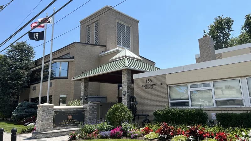 Mineola Village Hall on Washington Avenue, a tan brick building with flagpoles, a stone sign and a flower bed in front.
