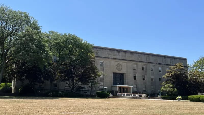 The long stone front of the Nassau County Court House seen across its lawn, framed by large trees.