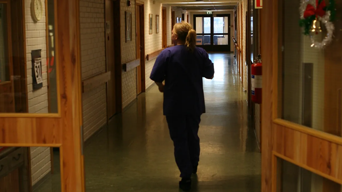 A staff member in scrubs walking down a long nursing-home corridor lined with handrails.