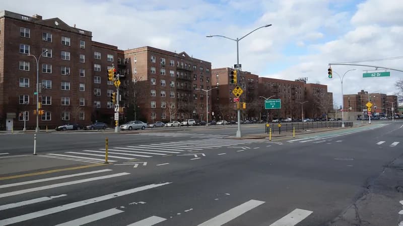 Wide marked crosswalks across Queens Boulevard at 62nd Drive in Rego Park, Queens, with traffic signals and apartment buildings behind.