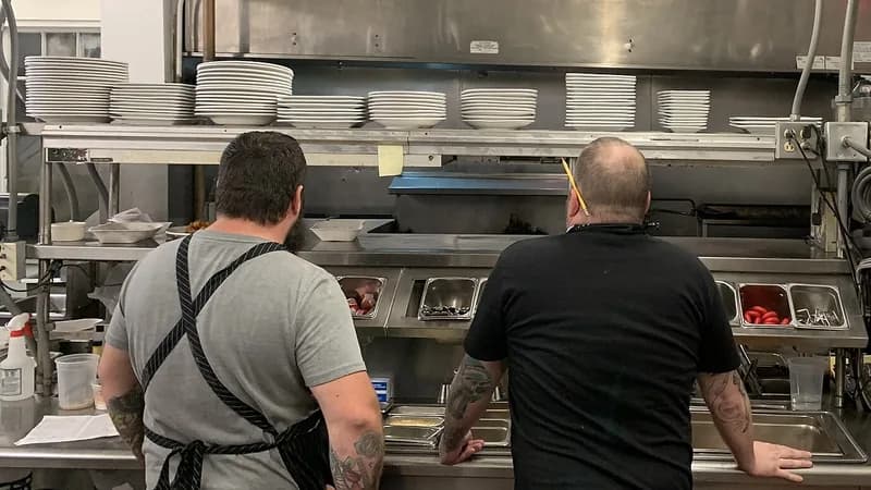 Two cooks seen from behind at the pass of a restaurant kitchen, with stacks of plates on the shelves.