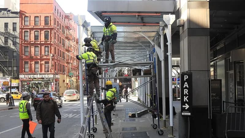 Construction workers in safety vests on ladders and scaffolding setting up a sidewalk shed on a Manhattan street.