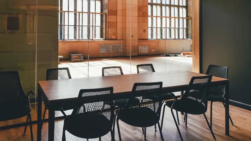 A small conference room with a wooden table and mesh chairs behind a glass wall, sunlit windows beyond.