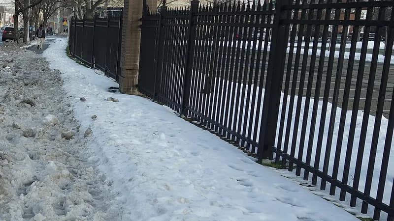 A snow-covered sidewalk with a trampled path beside an iron fence on Union Turnpike in Hillcrest, Queens.
