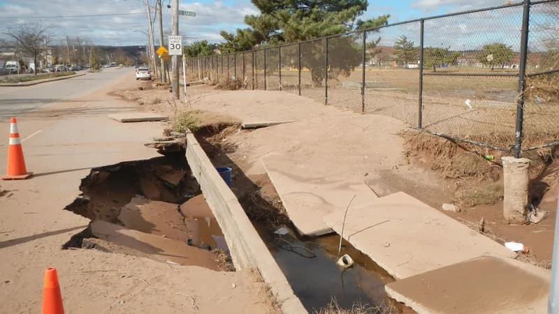 A collapsed and undermined concrete sidewalk beside a road on Crispo Way, Staten Island, marked off with orange traffic cones.